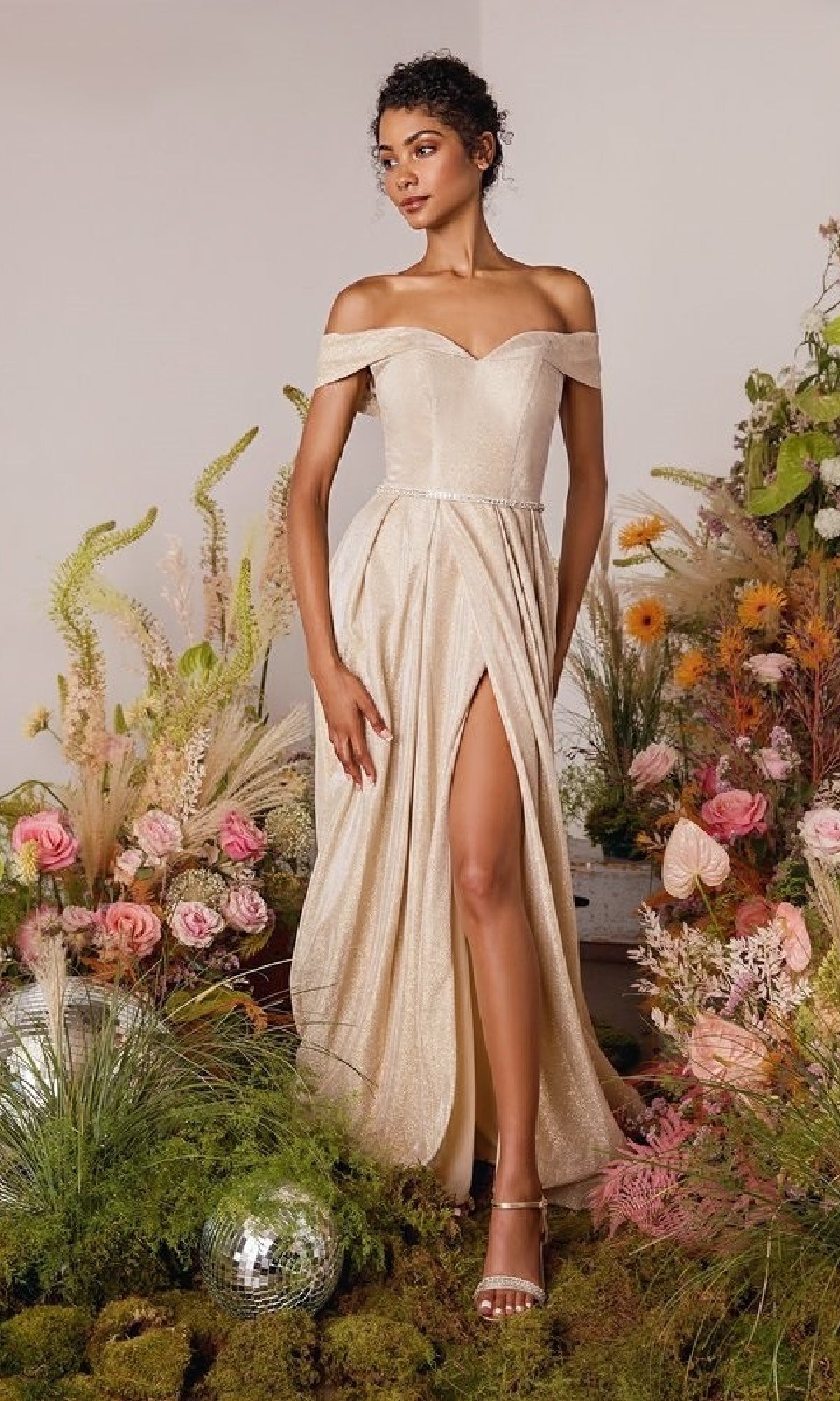 A woman in the Off-Shoulder Long Glitter Prom Dress: Eureka 9808, featuring an off-the-shoulder neckline and thigh-high slit, stands among flowers and greenery beside a disco ball.